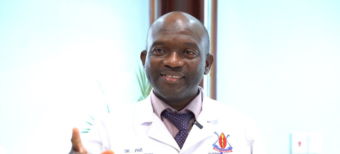 Daktari wa kiume aliyevaa koti jeupe lenye lebo ya jina na nembo ya hospitali anaelekeza kamera.