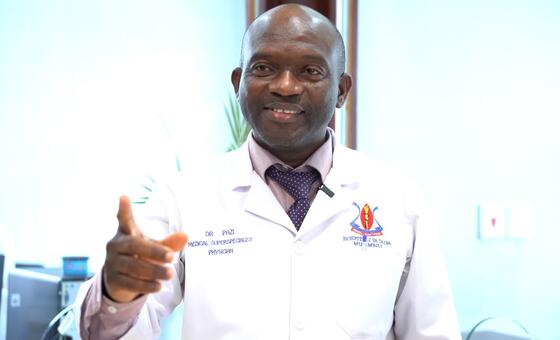 Daktari wa kiume aliyevaa koti jeupe lenye lebo ya jina na nembo ya hospitali anaelekeza kamera.