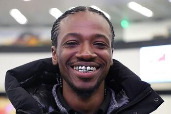Primer plano de un hombre afroamericano sonriente con el pelo trenzado, que lleva una chaqueta negra y una parrilla dental plateada.