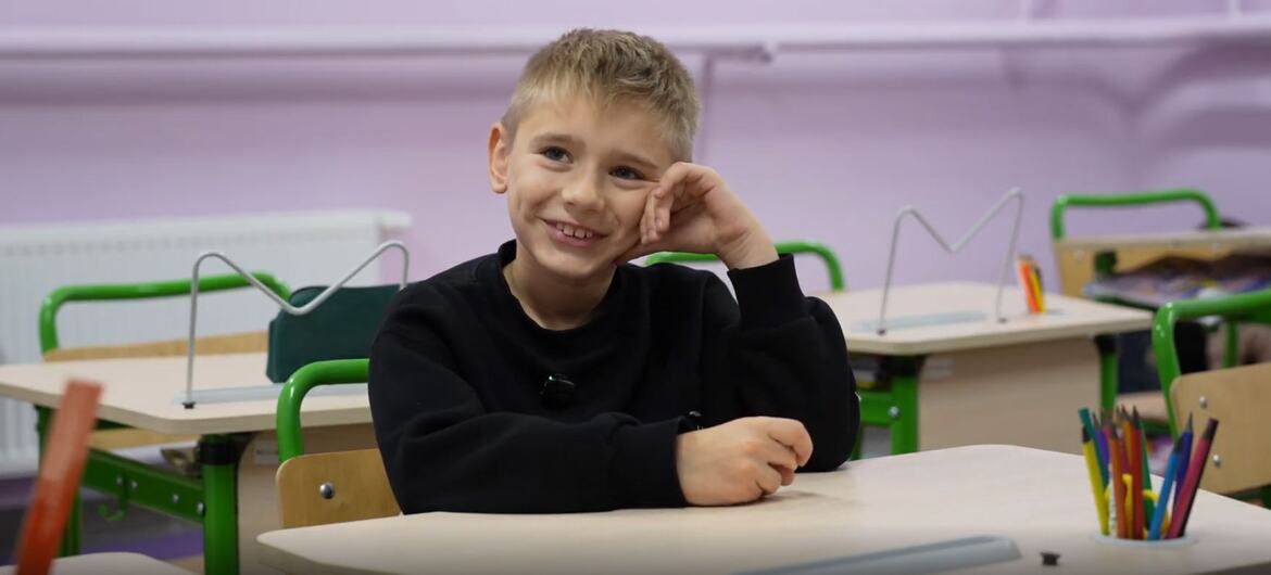A young boy sits at a school desk and smiles at the camera.