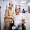 Une femme âgée se tient derrière un homme en fauteuil roulant, la jambe bandée, dans un couloir de réadaptation d'un centre médical.