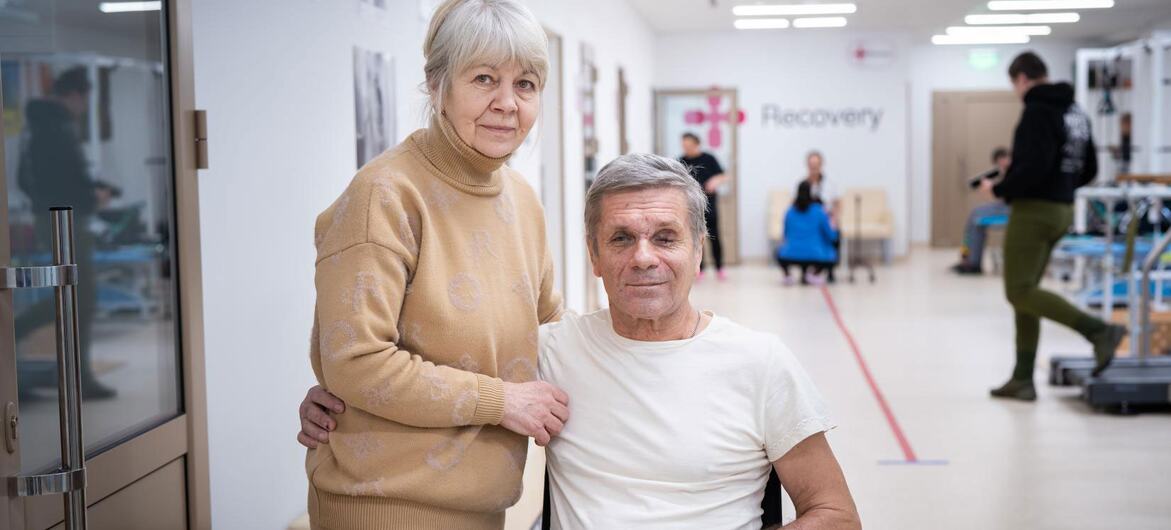 An elderly woman stands behind a man in a wheelchair with a bandaged leg in a medical center rehabilitation hallway.