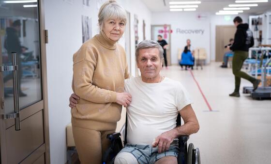 Une femme âgée se tient derrière un homme en fauteuil roulant, la jambe bandée, dans un couloir de réadaptation d'un centre médical.