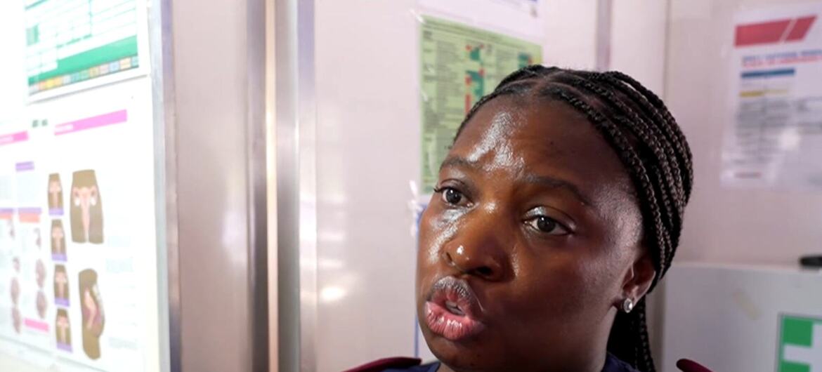 Mfanyakazi wa afya mwanamke mweusi mwenye nywele zilizosokotwa na sare ya bluu akiwa na epauleti za rangi ya hudhurungi akizungumza katika mazingira ya kliniki, huku mabango ya taarifa za kimatibabu yakiwa ukutani nyuma yake.