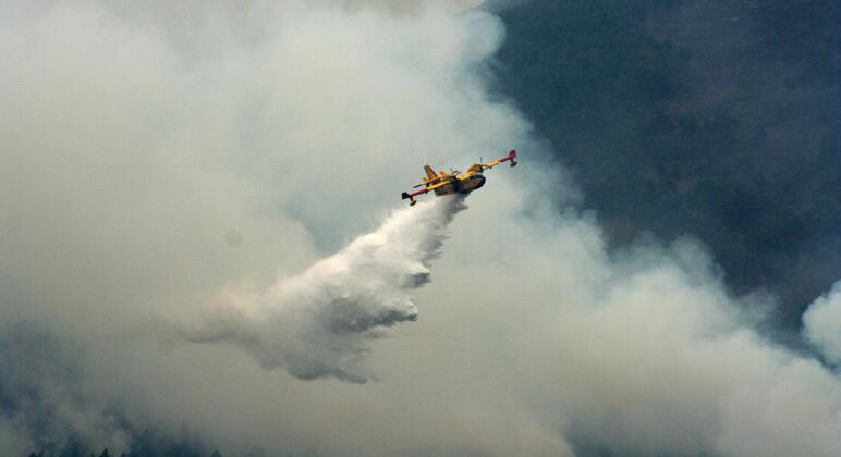 Un avión amarillo de extinción de incendios arroja un gran volumen de agua o retardante sobre un incendio forestal que produce mucho humo.