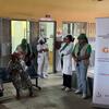 Um grupo de mulheres com uniformes verdes e profissionais de saúde estão na sala de espera de uma clínica em Moçambique, ao lado de uma faixa do projeto GRIS.