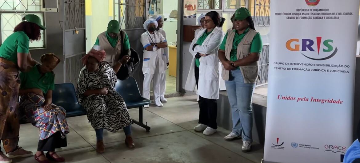Um grupo de mulheres com uniformes verdes e profissionais de saúde estão na sala de espera de uma clínica em Moçambique, ao lado de uma faixa do projeto GRIS.