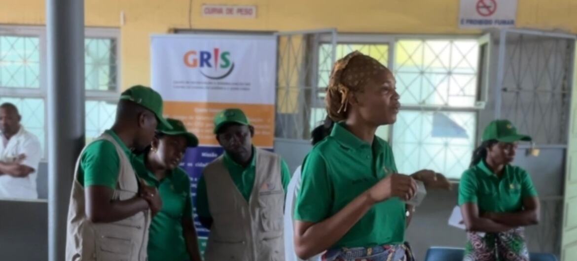 Uma mulher vestindo uma camisa polo verde e uma saia estampada conversa com um grupo de funcionários com uniformes combinando em tons de verde e bege em um evento do Unodc em Moçambique.