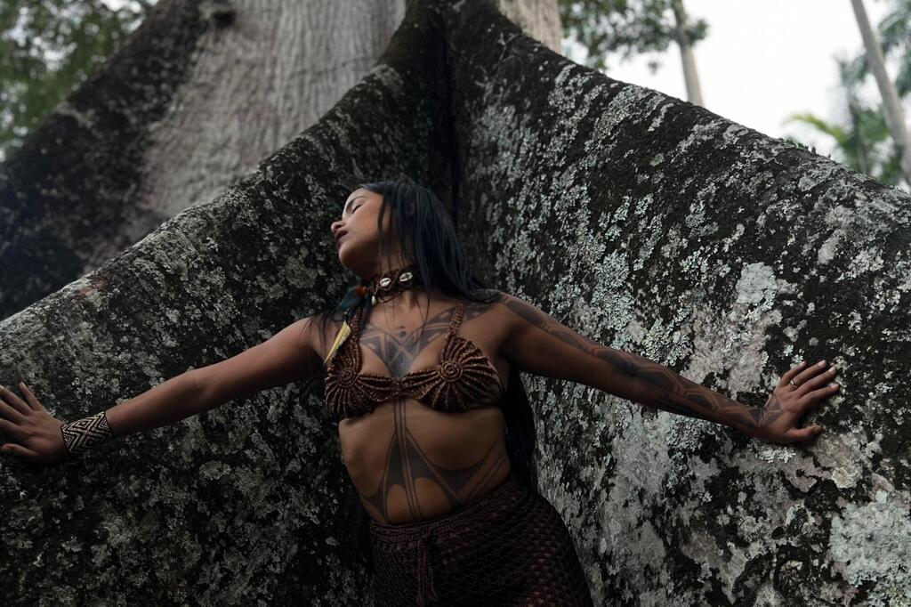 Une femme autochtone, vêtue de vêtements et portant des tatouages ​​traditionnels, se tient les bras écartés contre un grand arbre dans une forêt.