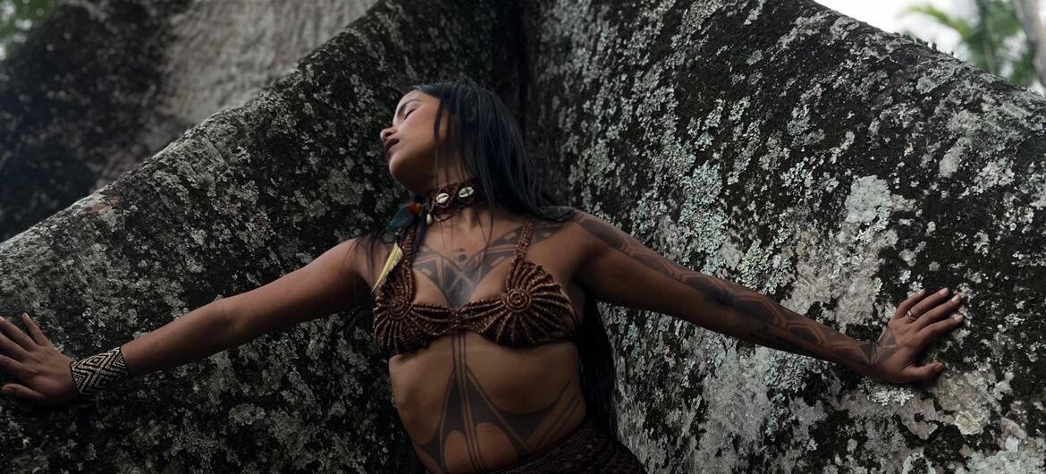 Uma mulher indígena com tatuagens e roupas tradicionais está de pé com os braços estendidos, encostada em uma grande árvore na floresta.