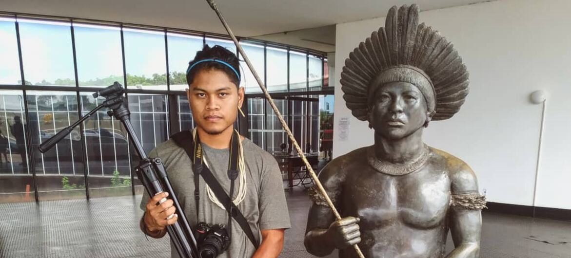 A Brazilian indigenous photographer holding a camera and tripod stands next to a bronze statue of an indigenous person with a feather headdress inside a museum.