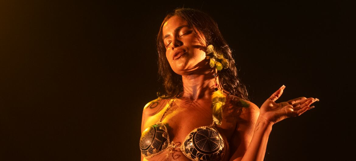 A female indigenous Brazilian singer with body paint and a flower in her hair performs on stage holding a microphone.