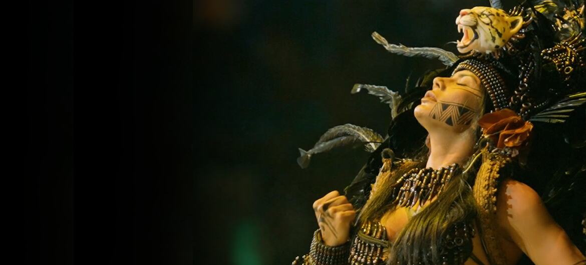 A female performer in elaborate indigenous-inspired carnival costume with a large feathered headdress and face paint performs on stage.