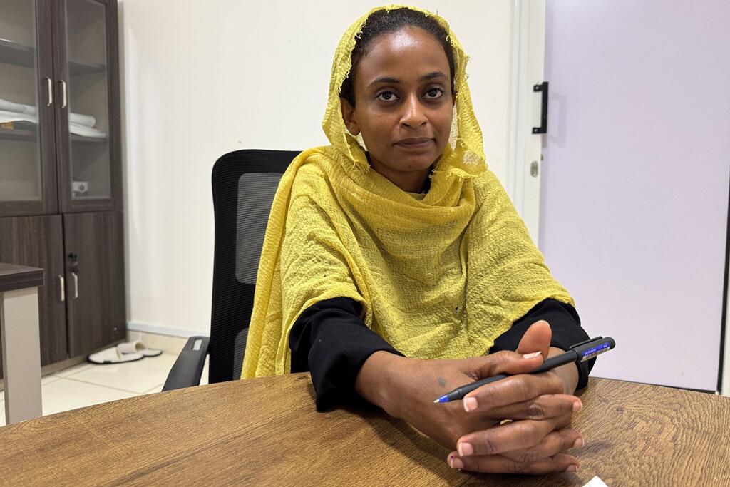 Une étudiante soudanaise portant un foulard jaune est assise à un bureau, un stylo à la main, et regarde droit dans l'objectif.