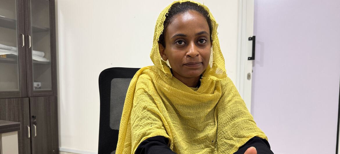 A Sudanese student wearing a yellow headscarf sits at a desk in an office, holding a pen and looking directly at the camera.