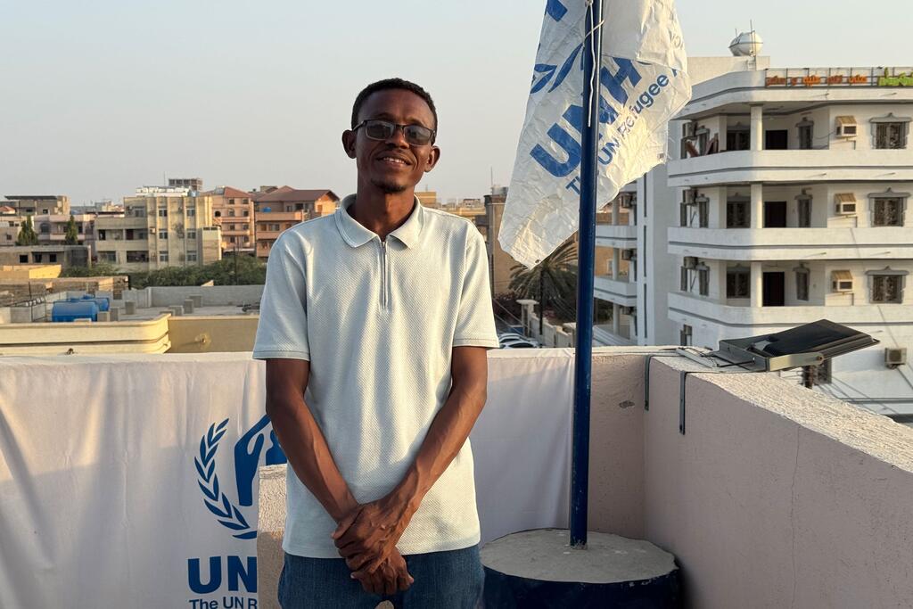 Un jeune étudiant souriant se tient sur un toit à côté d'un drapeau et d'une banderole du HCR au Soudan.