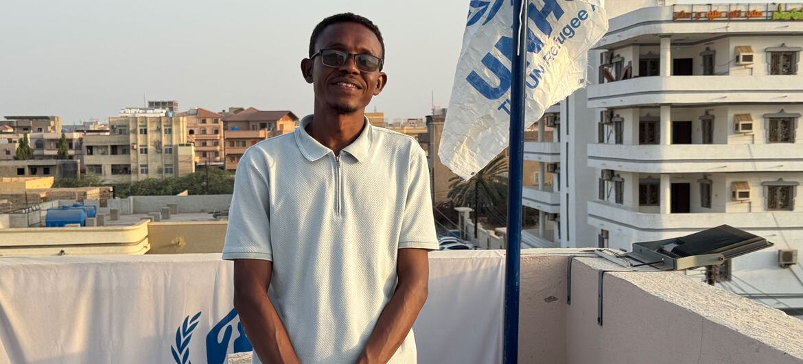 A smiling young male student stands on a rooftop next to a UNHCR flag and banner in Sudan.