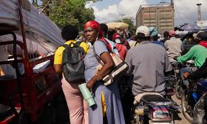 Une femme portant un foulard rouge se tient dans une rue haïtienne bondée, au milieu de motos et de camions.