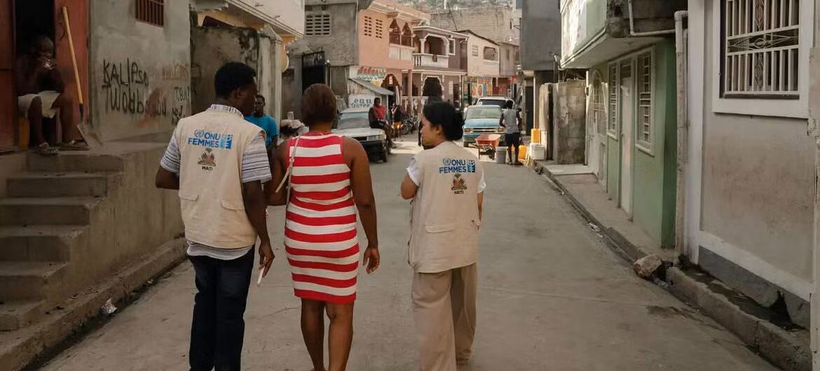 Deux employées ONU Femmes et une femme du quartier marchent dans une rue étroite d'un quartier haïtien.