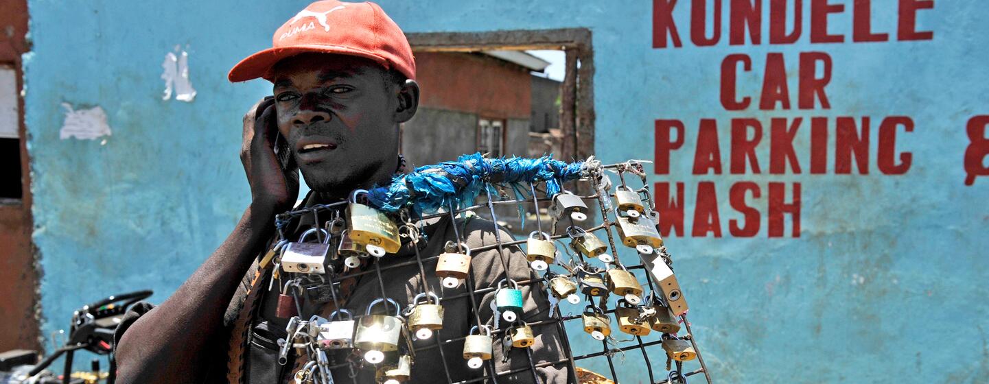 Un jeune homme noir portant une casquette rouge se tient dans une rue de Kisumu, au Kenya, vendant des cadenas tout en parlant au téléphone portable.