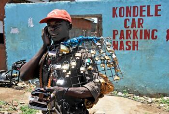 Um jovem negro usando um boné vermelho está na rua em Kisumu, no Quênia, vendendo cadeados enquanto fala ao celular.