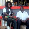 Uma mulher negra sorridente está sentada em um caixote de madeira ao lado de um homem negro em uma banca de engraxate ao ar livre em Moçambique.