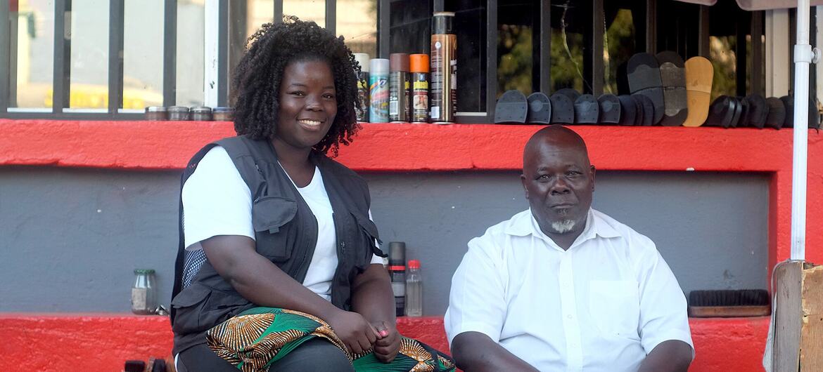 Uma mulher negra sorridente está sentada em um caixote de madeira ao lado de um homem negro em uma banca de engraxate ao ar livre em Moçambique.