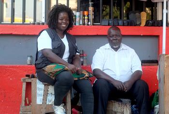 Uma mulher negra sorridente está sentada em um caixote de madeira ao lado de um homem negro em uma banca de engraxate ao ar livre em Moçambique.