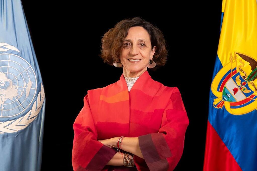 Una mujer de pelo castaño corto sonríe a la cámara, de pie entre una bandera de las Naciones Unidas y una bandera de Ecuador.
