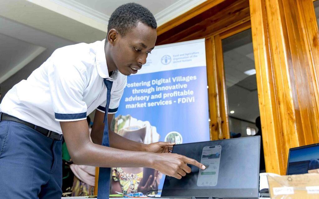 Un jeune homme noir, vêtu d'une chemise blanche et d'une cravate, désigne du doigt l'écran d'un ordinateur portable affichant l'interface d'une application mobile lors de l'événement Digital Villages Initiative.