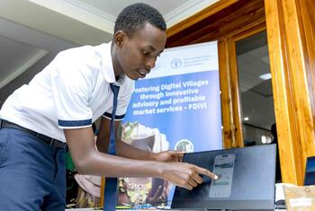 A young Black man in a white shirt and tie points to a laptop screen displaying a mobile app interface during the Digital Villages Initiative event.