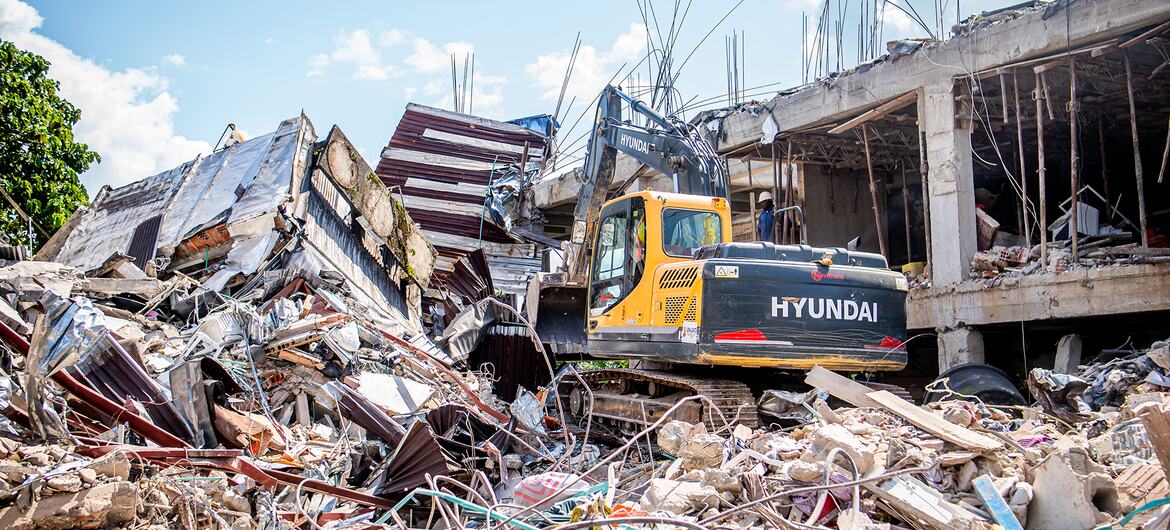 A yellow Hyundai excavator works on a large pile of rubble and destroyed buildings under a blue sky.