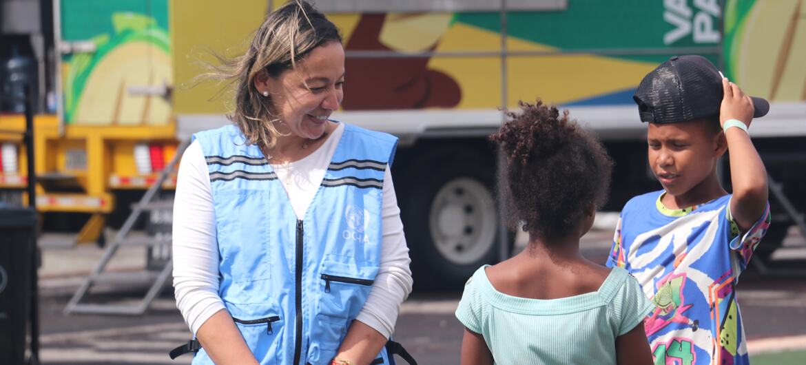 Una trabajadora de OCHA, vestida con un chaleco azul, sonríe mientras interactúa con dos niños durante un acto de ayuda humanitaria en Venezuela.