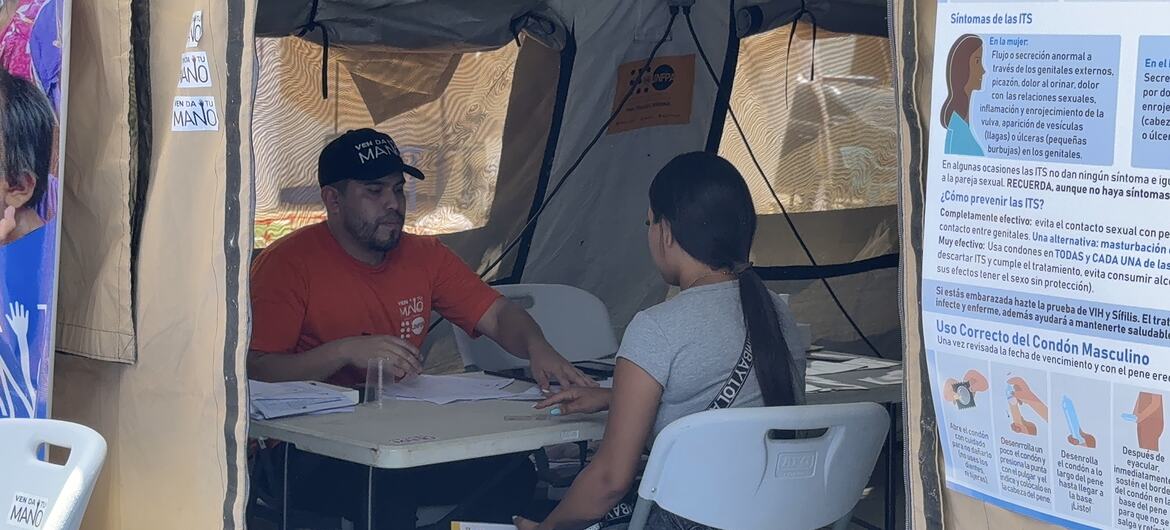 Un hombre con una camisa naranja y una mujer están sentados a una mesa dentro de una carpa durante una jornada de concienciación sobre salud. A la derecha se puede ver un cartel informativo sobre infecciones de transmisión sexual.