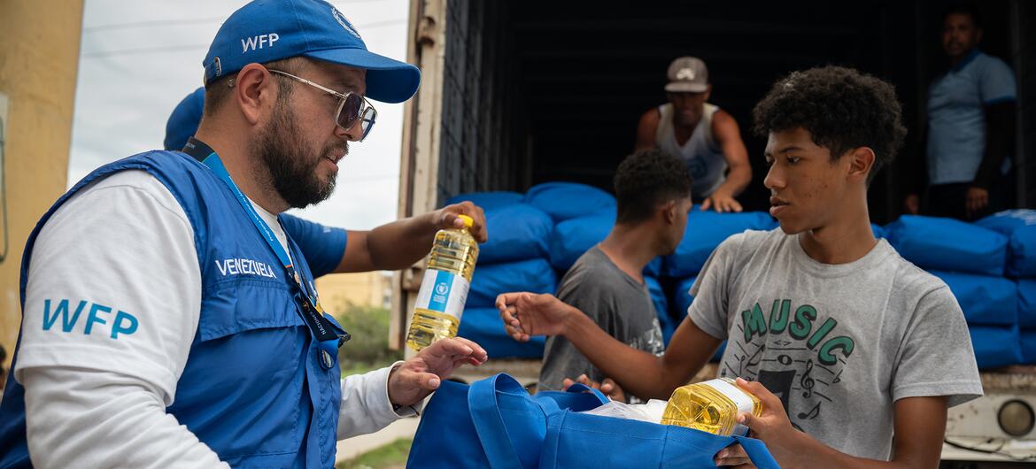 Un cooperante del PMA , vestido con un chaleco y una gorra azules, entrega botellas de aceite de cocina a un joven que recibe ayuda alimentaria en Venezuela.