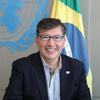 Igor Garafulic sorri em um retrato formal com as bandeiras da ONU e do Brasil ao fundo.