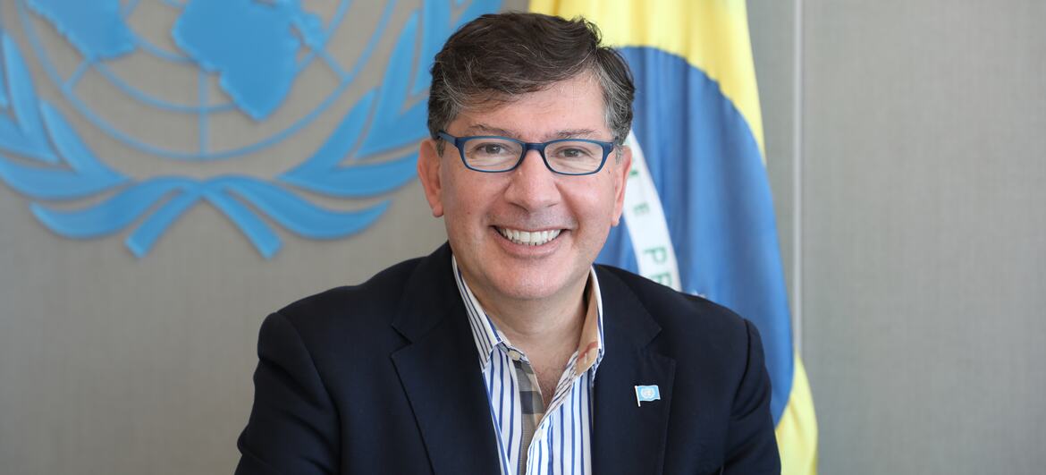 Igor Garafulic sorri em um retrato formal com as bandeiras da ONU e do Brasil ao fundo.