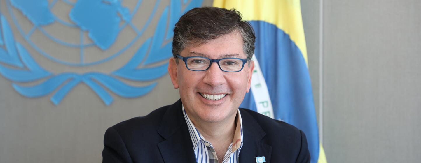 Igor Garafulic sorri em um retrato formal com as bandeiras da ONU e do Brasil ao fundo.