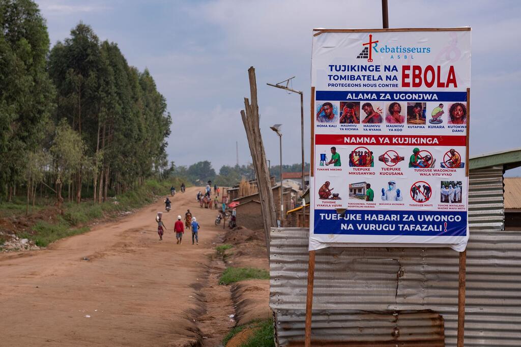 Bango la kuzuia Ebola limesimama kando ya barabara ya vumbi katika Mkoa wa Ituri, DRC, huku watu wakitembea kando ya njia.