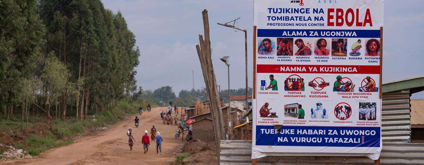 Une affiche de prévention contre Ebola est dressée au bord d'un chemin de terre dans la province d'Ituri, en RDC, tandis que des personnes empruntent ce chemin.