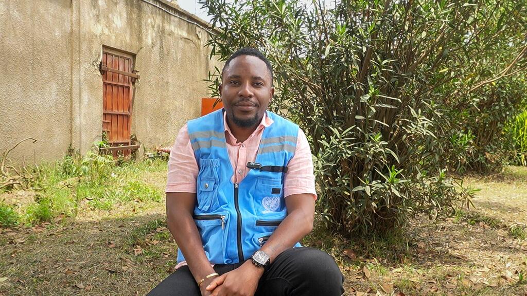 Hubert Amani, un homme noir aux cheveux courts et à la barbiche, portant un gilet bleu de l'OCHA des ONU sur une chemise rose, est assis par terre à l'extérieur, devant un bâtiment et des buissons.
