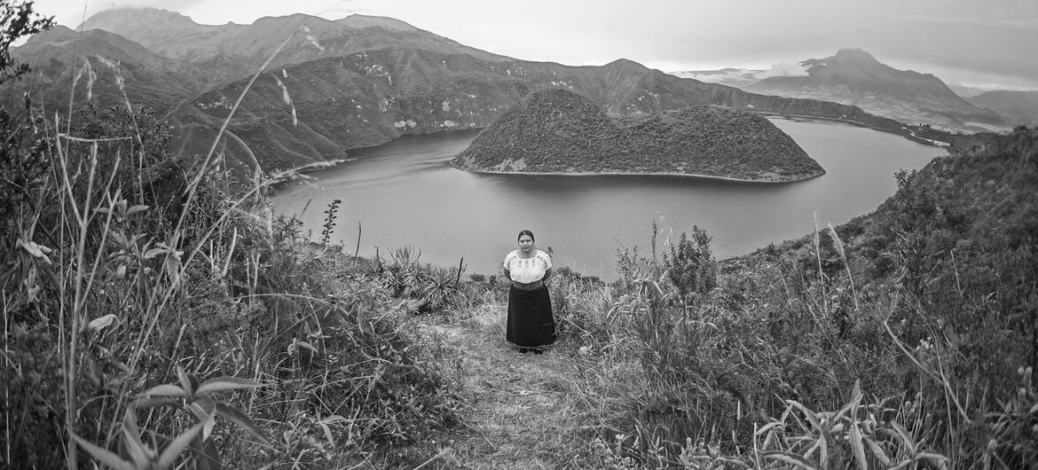 Una mujer vestida con ropa tradicional ecuatoriana se encuentra de pie en la cima de una colina cubierta de hierba, con vistas a un gran lago con una isla y las montañas que lo rodean.