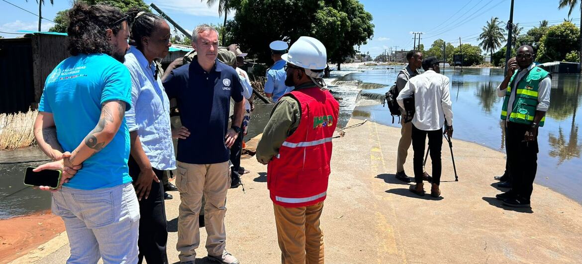 Um grupo de funcionários e trabalhadores humanitários, incluindo Sergio Vieira de Mello, está em uma estrada asfaltada ao lado de uma área alagada em um ambiente tropical.