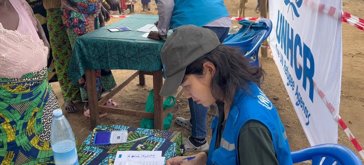 一名身穿蓝色背心、头戴帽子的UNHCR女工作人员在户外登记处填写表格。