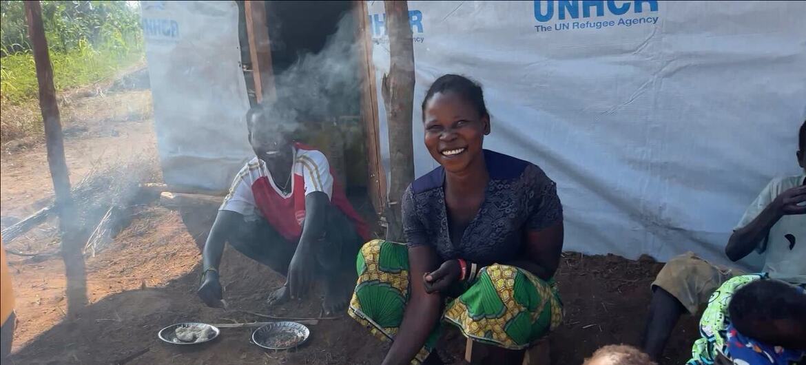 一位面带微笑的黑人妇女和一个小男孩在UNHCR帐篷庇护所前用明火做饭。