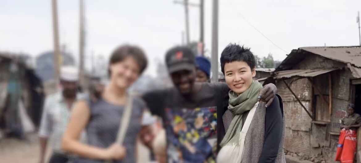 一位留着短发、系着绿色围巾的亚洲女性面带微笑地站在一位戴着帽子的非洲男性旁边。他们站在一条街道上，背景是一些简朴的建筑。
