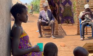 A young African girl in a colorful dress leans against a wall holding a cup while two older men sit on chairs in the background of a rural village.