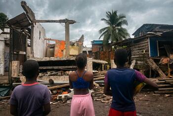 Three children stand looking at damaged wooden and concrete buildings in a neighborhood after an earthquake.