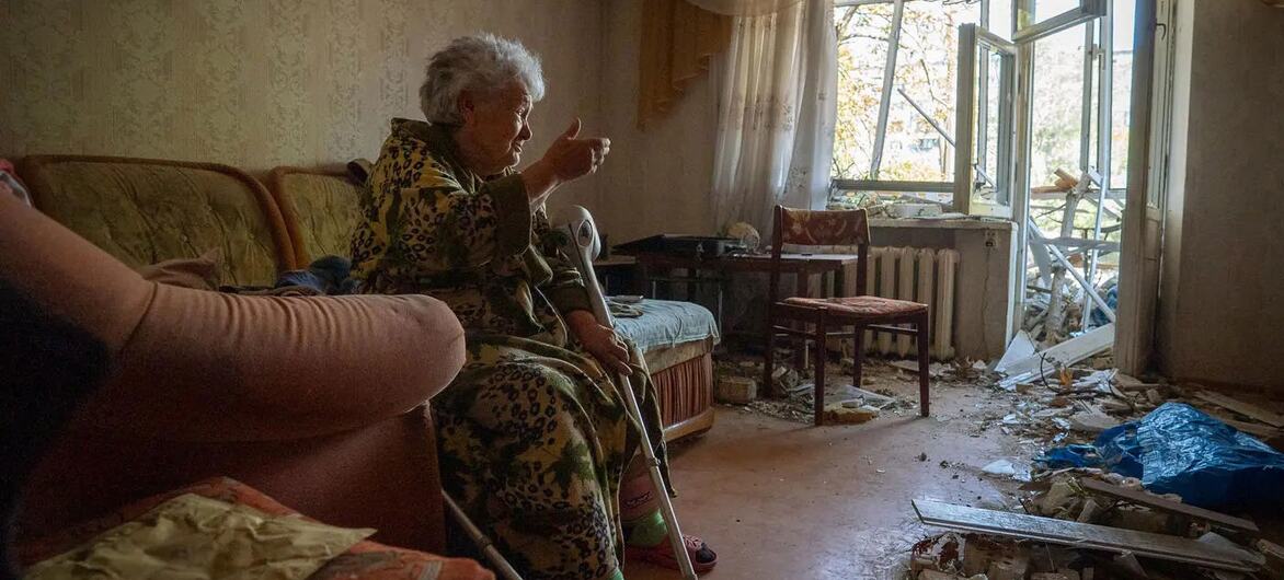 An elderly woman sits in her damaged apartment surrounded by debris and a broken window in Ukraine.