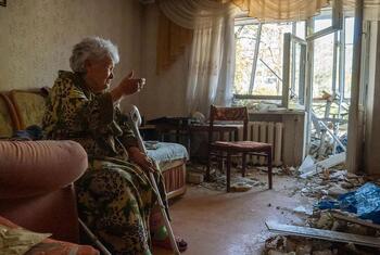 An elderly woman sits in her damaged apartment surrounded by debris and a broken window in Ukraine.